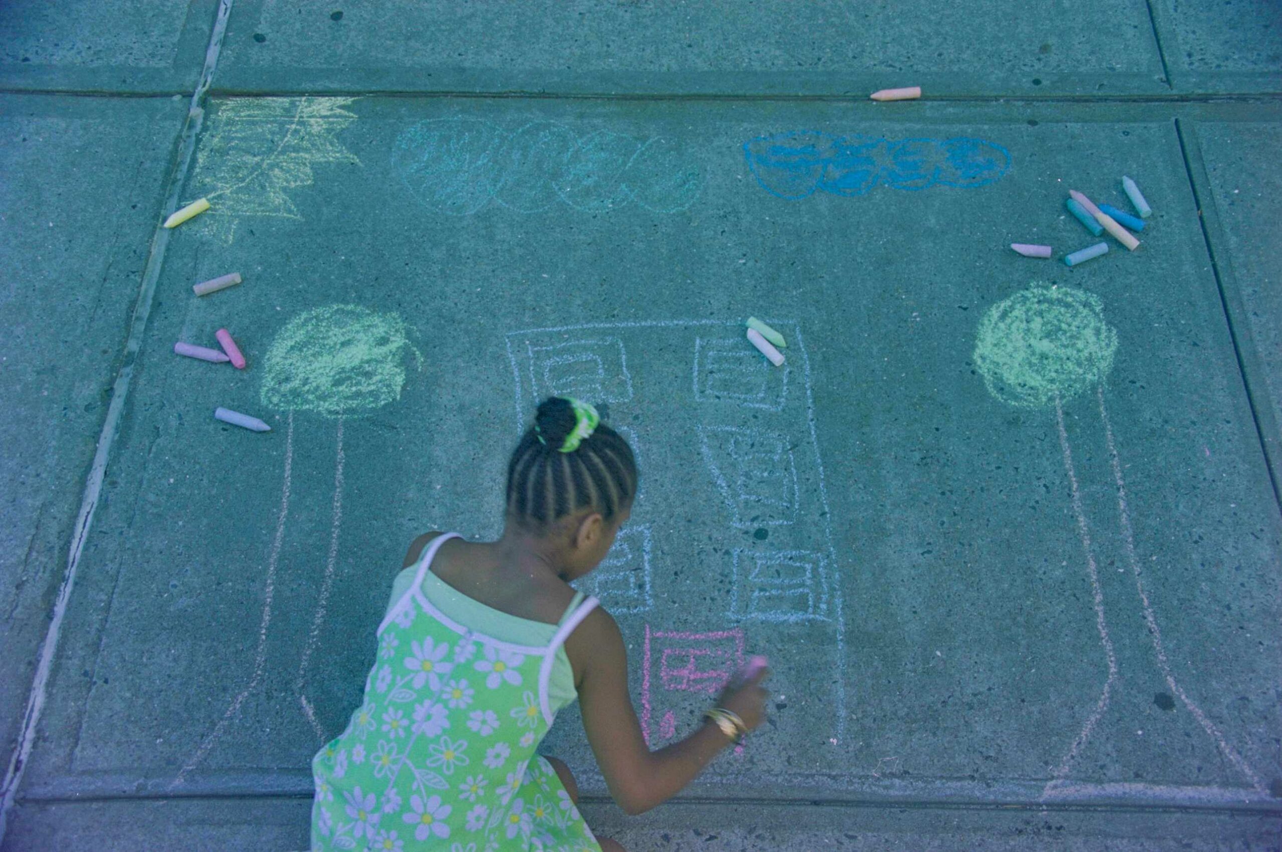 A young girl in a colorful flowered dress drawing an image of a home and trees